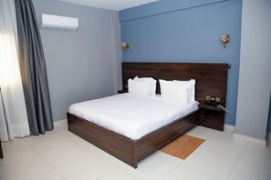Quarto de hotel com uma cama grande, mesa de cabeceira, ar condicionado fixado na parede e janela com cortinas.
