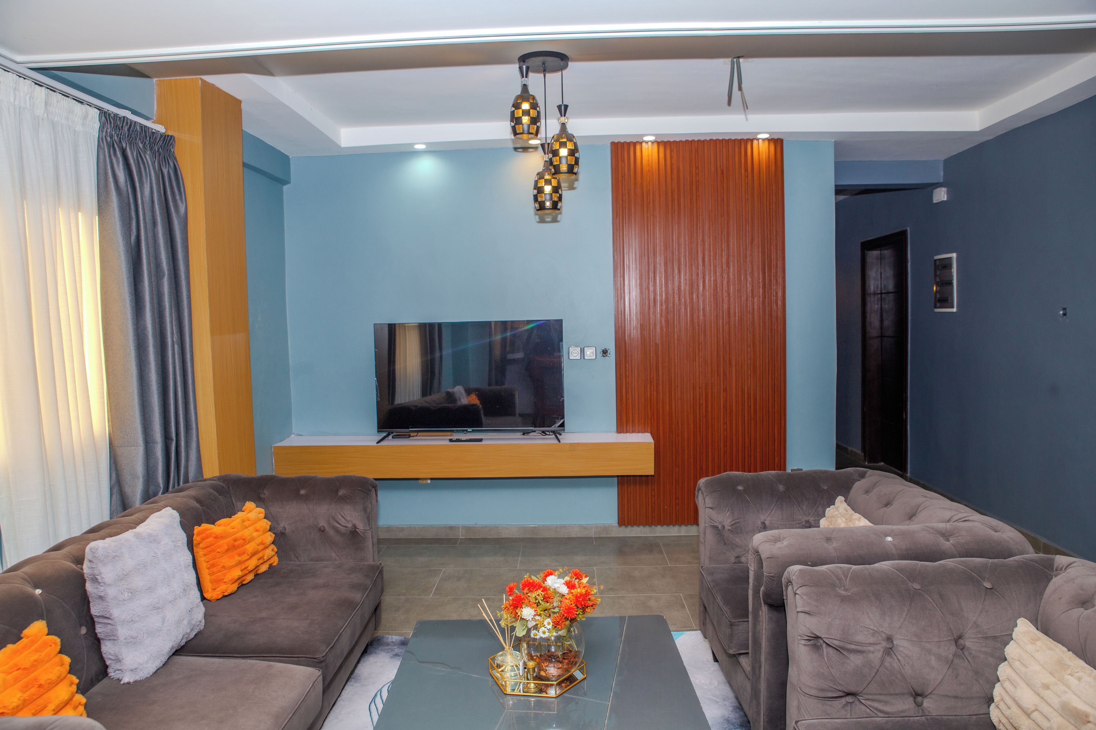 Uma sala de estar com dois sofás, uma mesa de centro com um vaso de flores, uma TV de tela plana sobre um suporte de madeira e uma parede revestida de madeira.