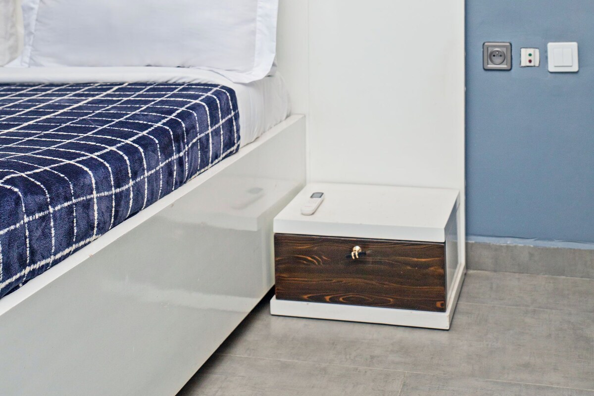 A bed with a wooden headboard, white bedding, and a blue checkered blanket. There is a bedside table with a drawer, a wall-mounted light, and a power outlet.