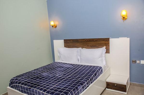 Un lit simple avec une couvre-lit à carreaux bleus et blancs, deux appliques murales et une tête de lit en bois.