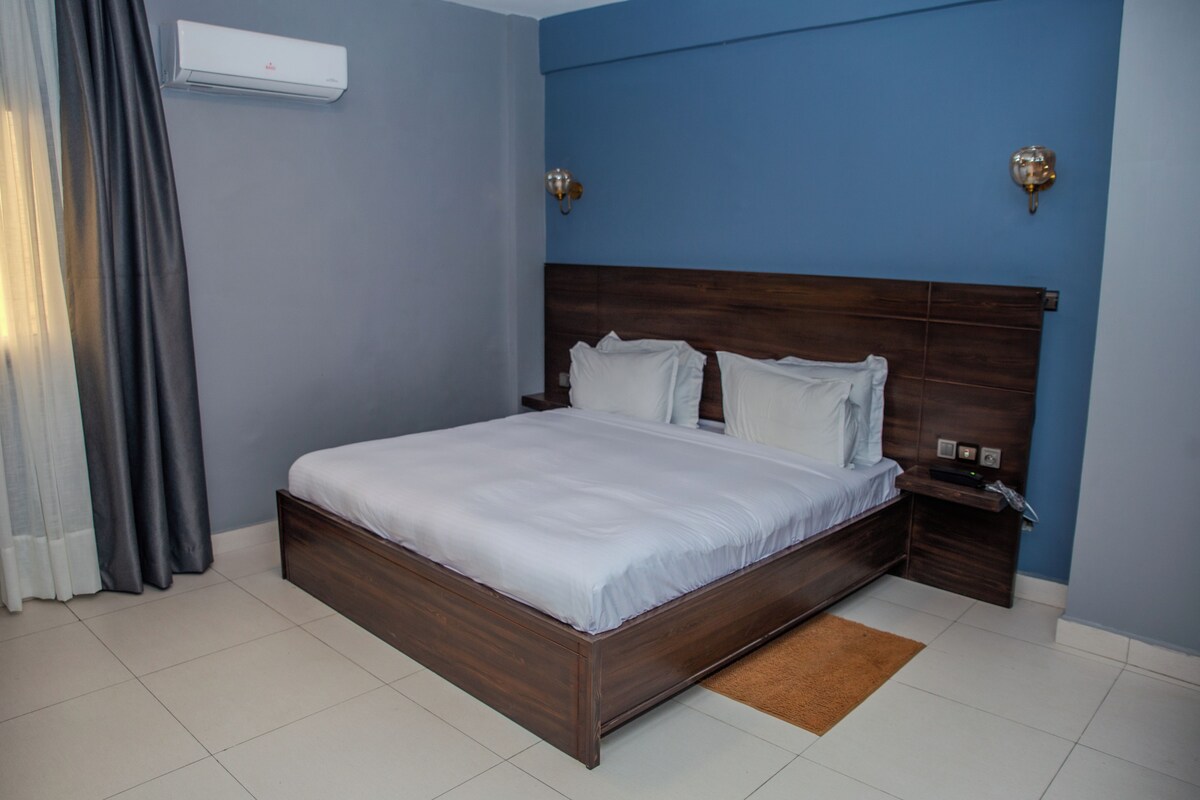 A hotel room with a wooden bed, white bedding, a brown rug, a nightstand, and a wall-mounted air conditioner.