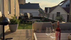An outdoor patio with a table, two wine glasses, a bottle of wine, a grill, and lounge chairs. - Apartment with a large terrace and a view of the Black Forest (Willstätt)