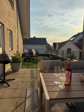 Eine Terrasse im Freien mit Tisch, zwei Weingläsern, einer Weinflasche, einem Grill und Lounge-Stühlen.