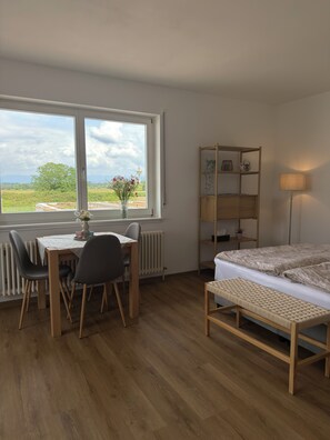 Ein Zimmer mit Holzboden, einem Bett mit weißer Matratze und beiger Bettwäsche, einem Holztisch mit Stuhl, einer Heizung und einem Fenster mit Blick auf Grünflächen und einen Turm.