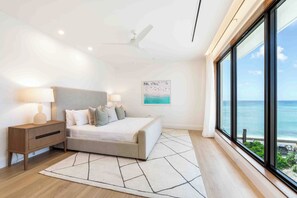 A modern bedroom with a bed, bedside tables, a ceiling fan, a large window with a view of the ocean, and a geometric rug. - The Sands 5: 7Mile Beach Condo w/Rooftop Pool & Beach Deck (West Bay)