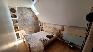 A bedroom with a sloped ceiling, a bed with a wooden headboard, a bedside lamp, and a shelf with decorative items. - Charming French cottage/charming country house (Le Plessis-Dorin)