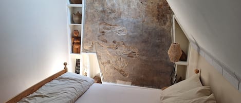 Un dormitorio con techo inclinado, cama con ropa de cama blanca, estante con objetos decorativos y pared texturizada.