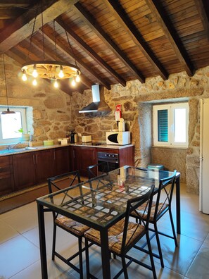 A rustic kitchen with stone walls, wooden beams, and a glass dining table. - Oak House - A Retreat in Caramulo (São João do Monte - Vale do Lobo)