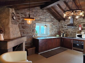 A rustic kitchen with stone walls, wooden beams, a built-in oven, and a hanging light fixture. - Oak House - A Retreat in Caramulo (São João do Monte - Vale do Lobo)
