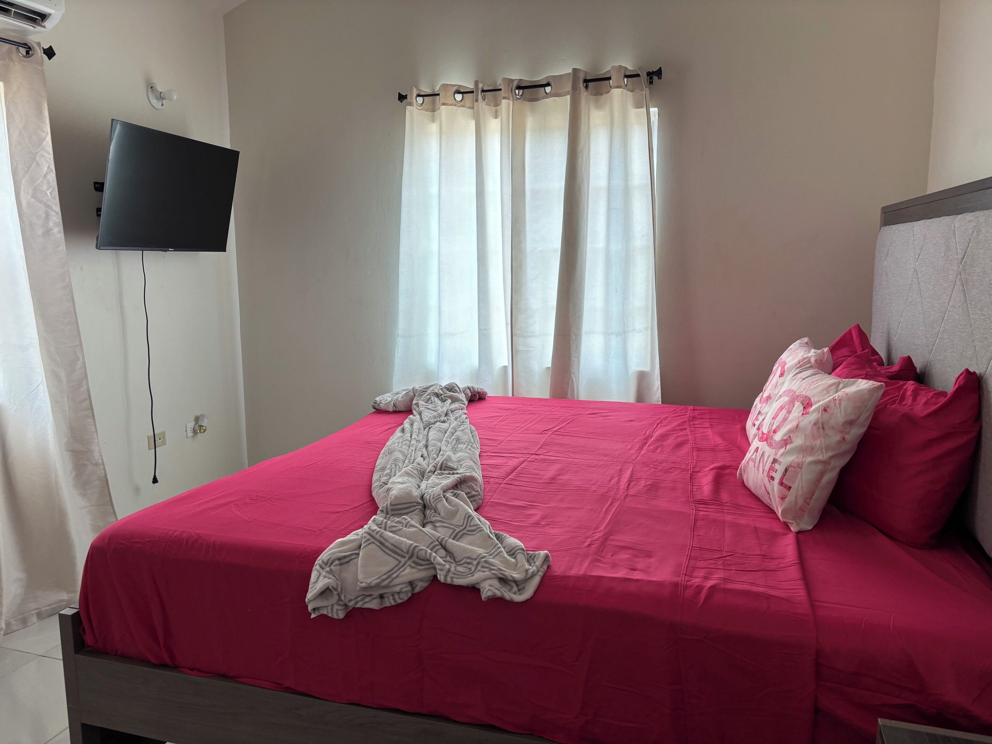 A bed with a pink bedspread, a blanket, and pillows, a flat-screen TV mounted on the wall, and a window with curtains.