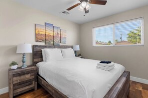A bedroom with a wooden bed, white bedding, a ceiling fan, a window with blinds, and a nightstand with a lamp and a plant. - Scottsdale Montecito Retreat | Pool, Hot Tub, Putting Green & Games 🌵⛳ (Scottsdale)