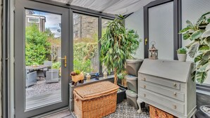Habitación con puerta de vidrio corredera que da a un espacio exterior, una cesta de mimbre, un taburete pequeño de madera, un aparador gris y una planta en maceta.