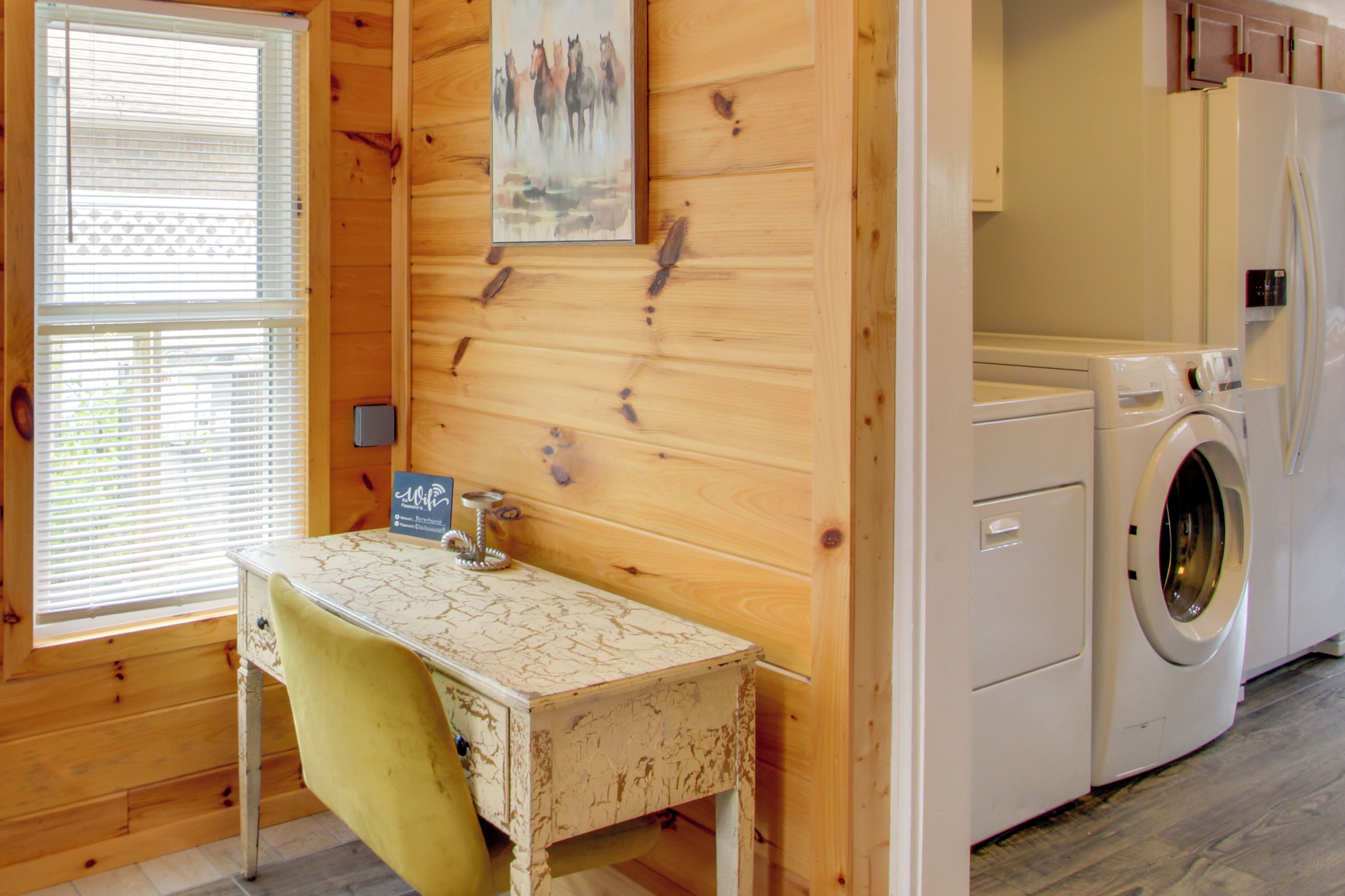 A room with wooden walls, a washing machine, a table with a chair, and a window with blinds.