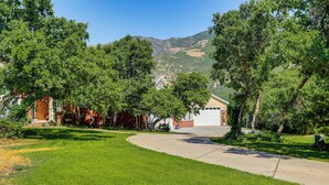 A residential driveway leading to a house with a garage, surrounded by lush green trees and a hilly landscape in the background. - Full Kitchen! King Suite Near Snowbasin (Layton)