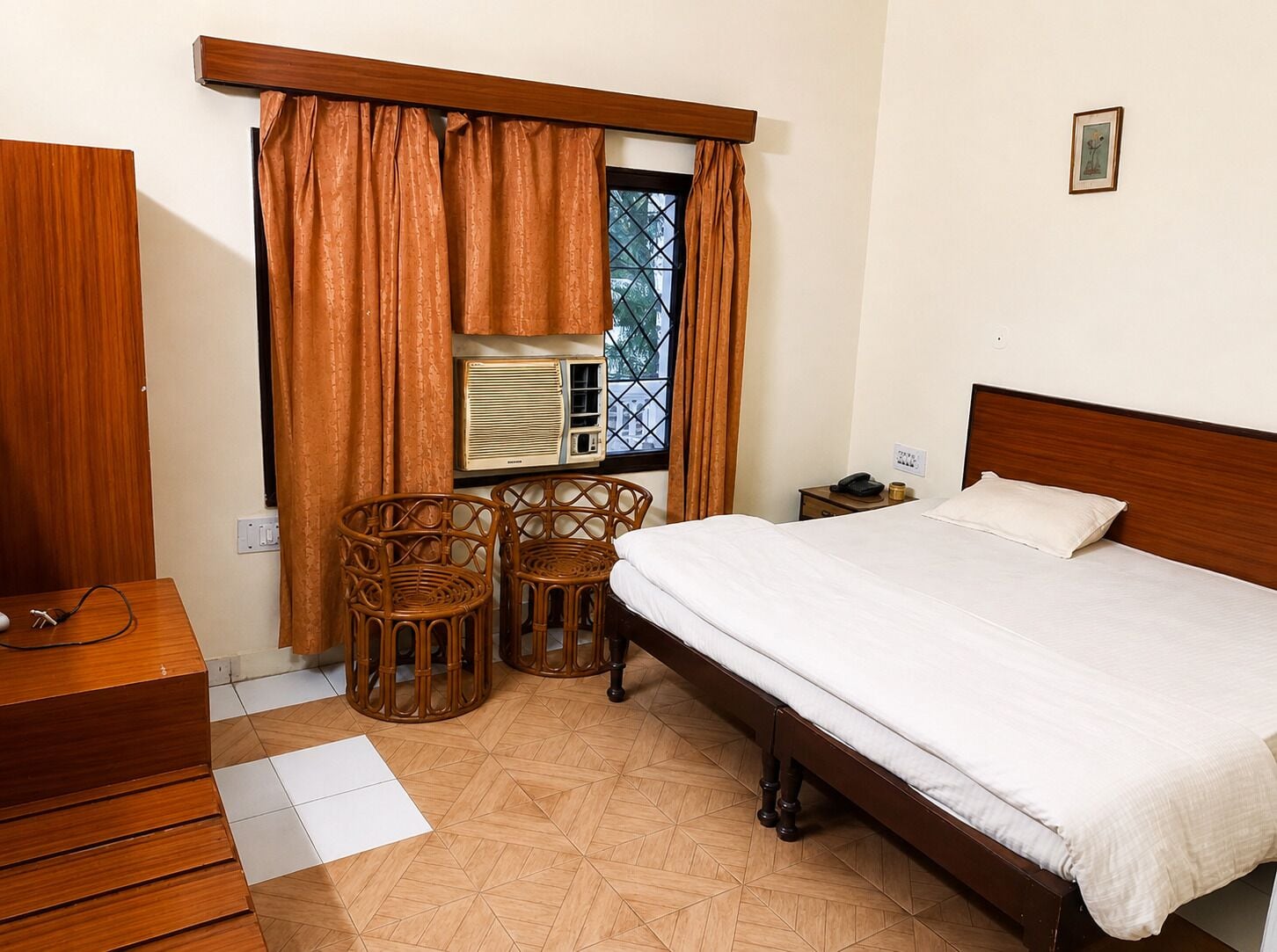 Habitación de hotel con cama, dos sillones de mimbre, una mesita de noche, un aparador, una ventana con cortinas y un radiador.