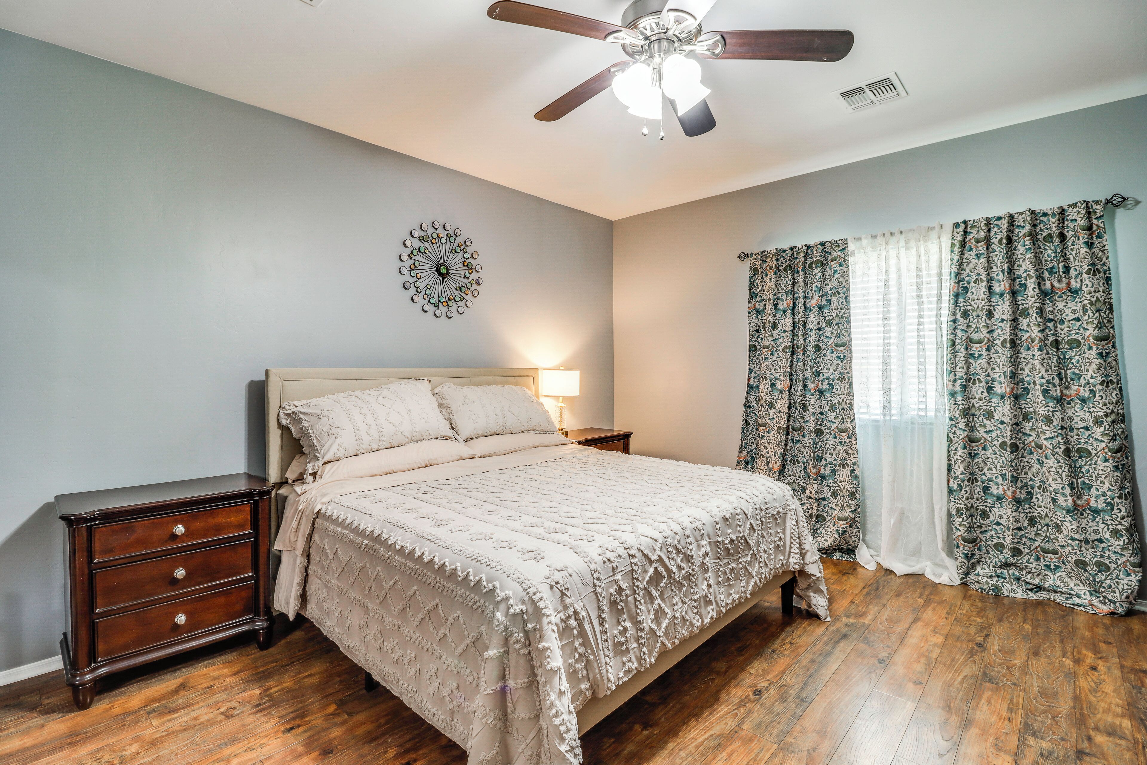 A bedroom with a bed, nightstand, dresser, ceiling fan, and patterned curtains.