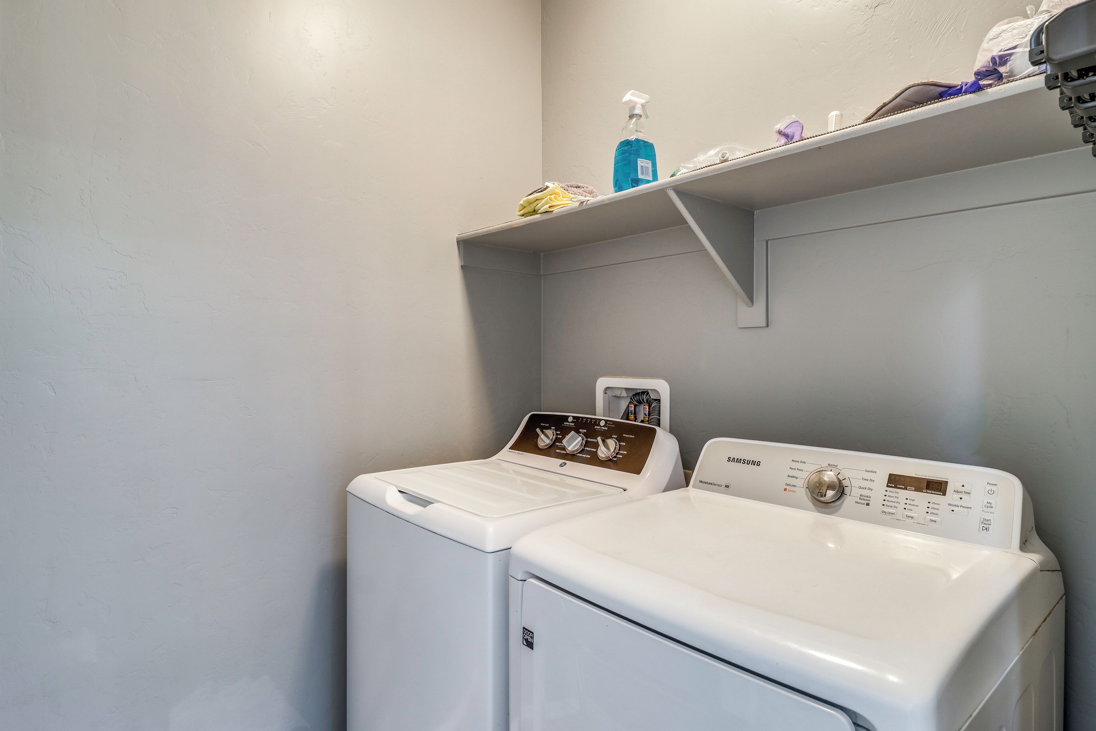 A laundry area with a front-loading washer and dryer, a shelf with cleaning supplies, and a wall-mounted control panel.