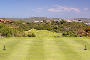 Ein Golfplatz mit blauem Himmel, einem gepflegten Grün und einem Blick auf Wohnhäuser im Hintergrund.