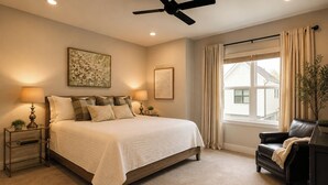 Ein Schlafzimmer mit Bett, Nachttischen, Schreibtisch, Sessel, Deckenventilator und einem Fenster mit Vorhängen.