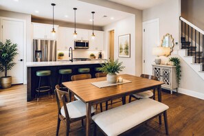 A modern kitchen with stainless steel appliances, a dining area with wooden furniture, and a staircase with a railing. - Magnolia Manor at College Station & Texas A&M (College Station)