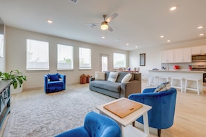 A spacious living room with a grey sofa, blue armchairs, a wooden coffee table, and a kitchen area with white cabinets and a red appliance. - Fenced Yard! Family Getaway, Near Texas Ski Ranch (San Marcos)
