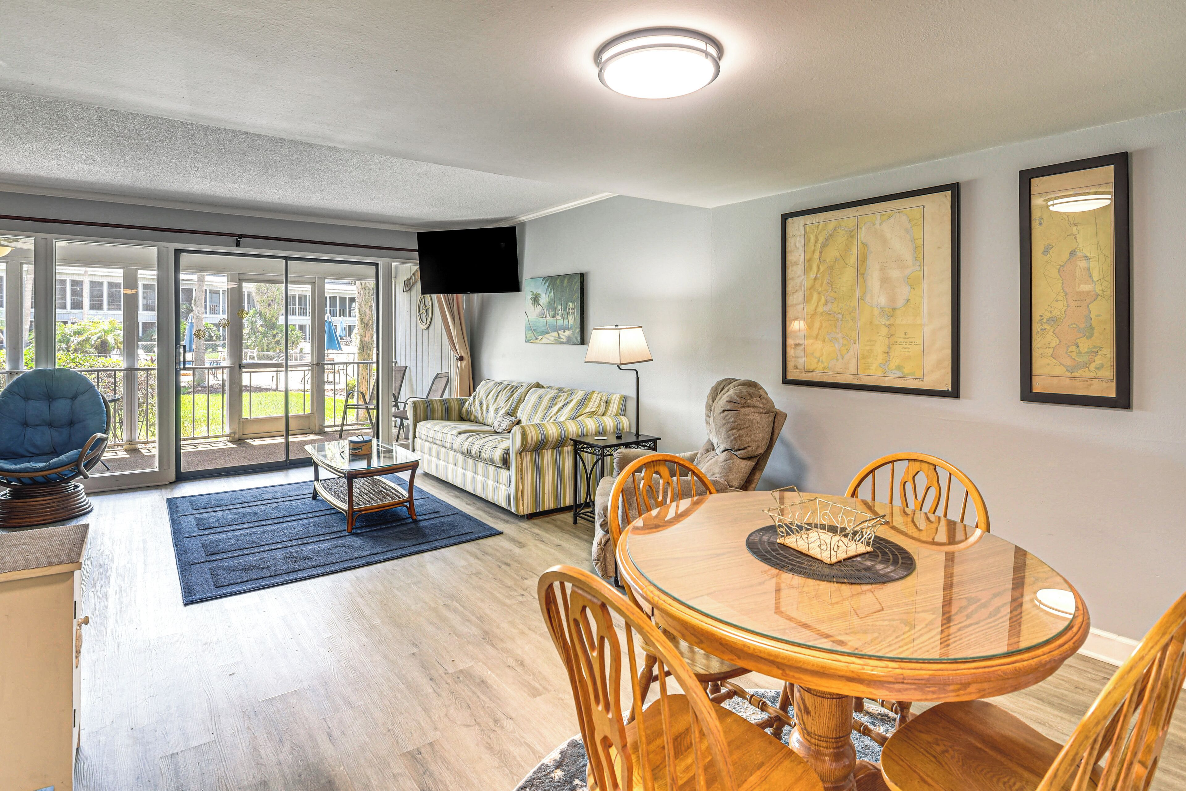 A spacious living room with a glass door leading to a balcony, a round dining table with chairs, a sofa, and a flat-screen TV mounted on the wall.