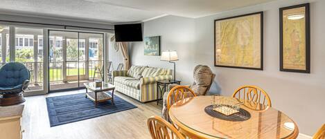 A spacious living room with a glass door leading to a balcony, a round dining table with chairs, a sofa, and a flat-screen TV mounted on the wall.