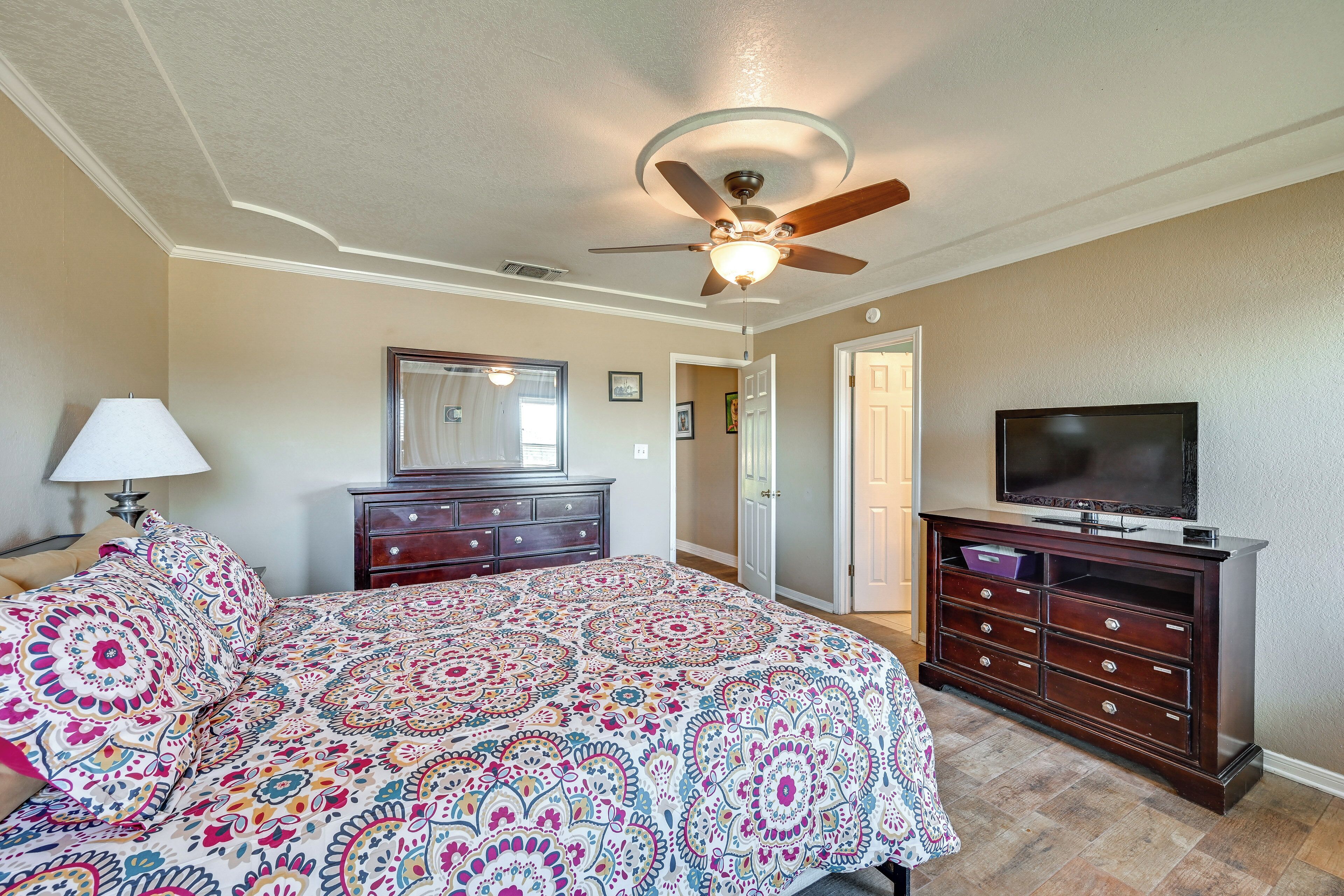 A bedroom with a bed, dresser, television, ceiling fan, and a door leading to another room.