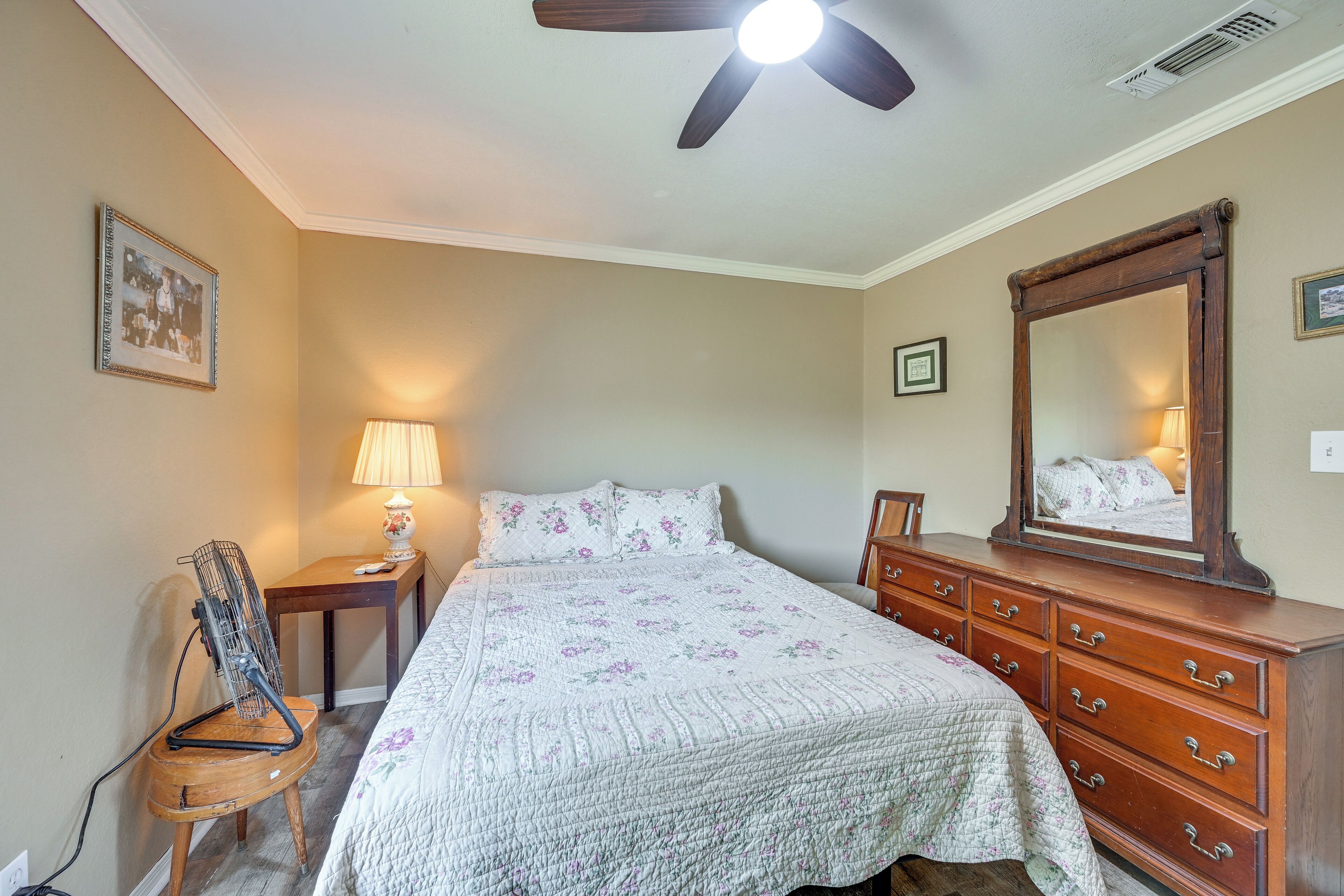 Ein Schlafzimmer mit Bett, Kommode, Spiegel, Lampe, Stuhl und Deckenventilator.
