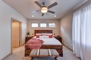 A bedroom with a bed, bedside tables, a bench, a ceiling fan, and three windows.