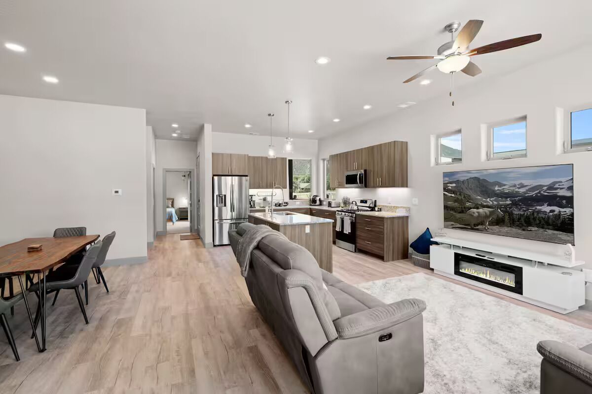 A modern kitchen with a central island, a dining area with a long table, and a living room with a large flat-screen TV.