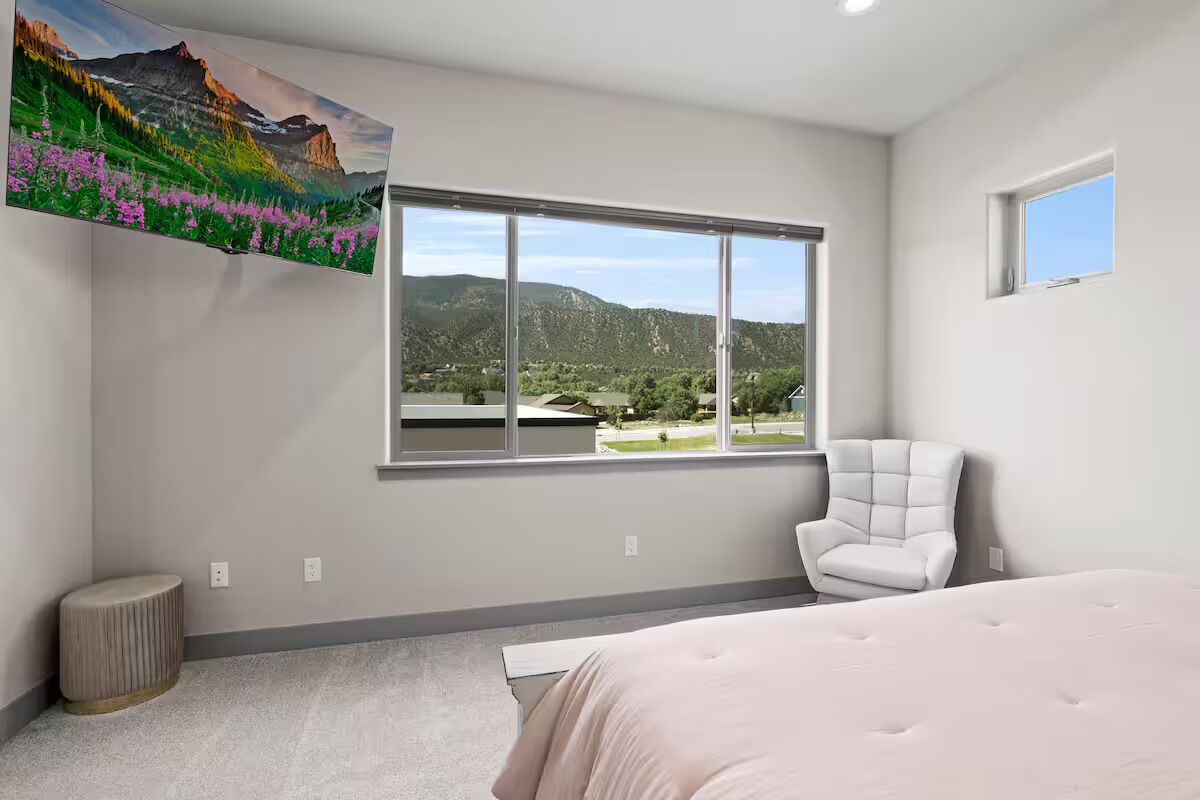 A bedroom with a large window, a bed, a chair, and a painting of a mountain landscape.