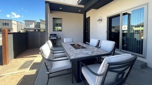 Um pátio externo com uma mesa retangular e seis cadeiras, uma churrasqueira e uma porta de vidro deslizante.