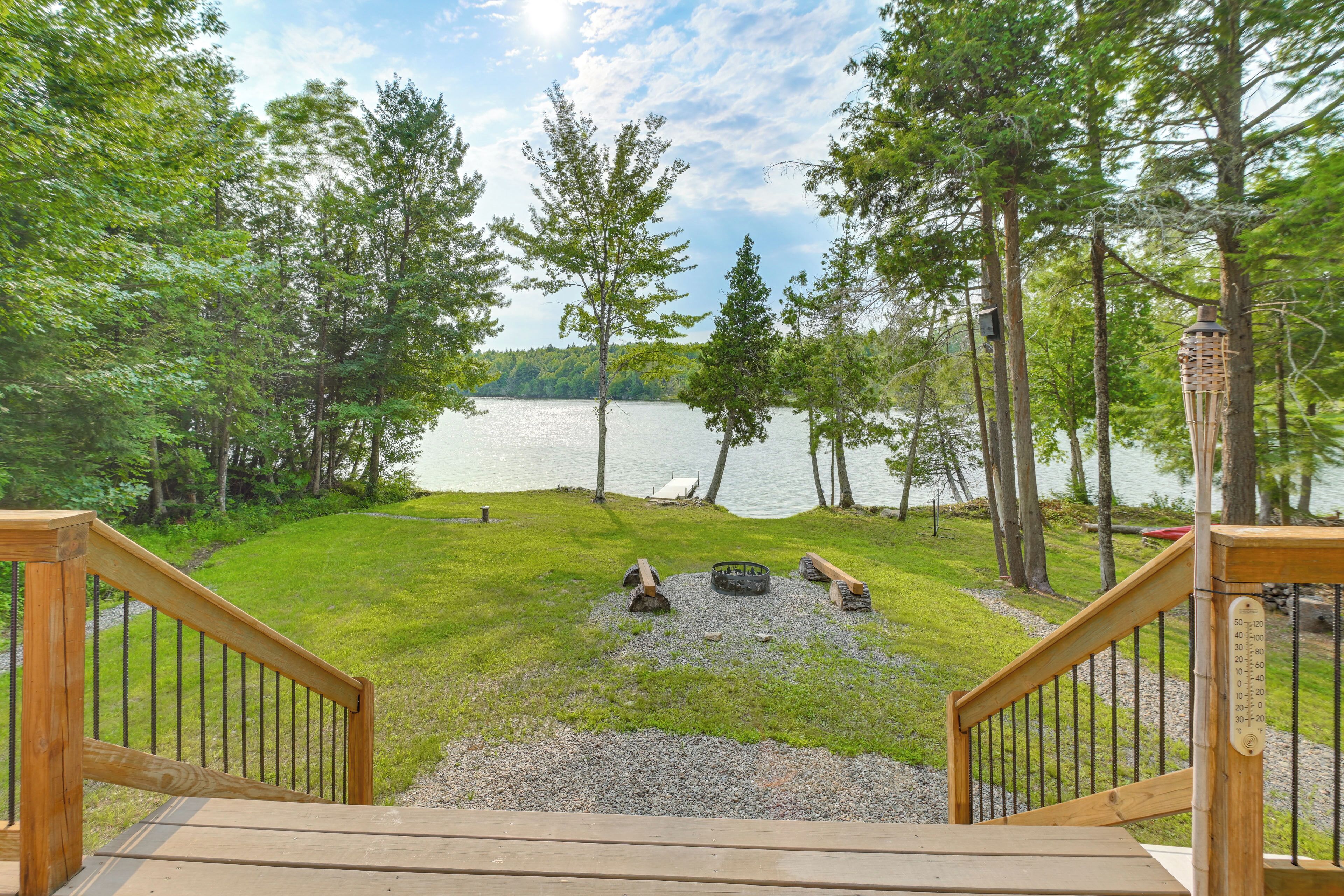 A wooden deck leading to a lakeside view with a fire pit and a thermometer.