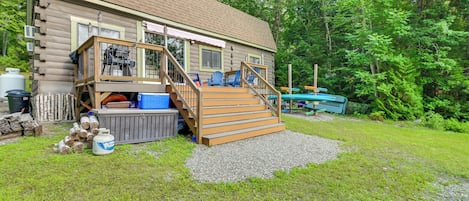 A log cabin with a deck, outdoor seating, a hot tub, and a gravel driveway.