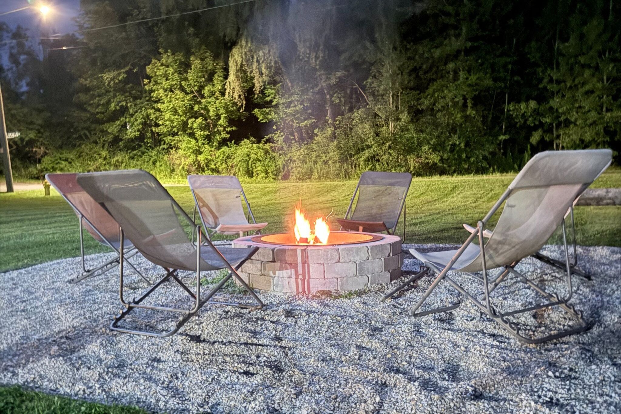 A fire pit surrounded by four chairs in a gravel area with a grassy field and trees in the background.