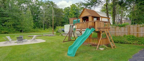 A wooden play structure with a slide, surrounded by a grassy yard and a wooden fence.