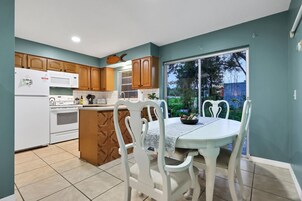A kitchen with wooden cabinets, a white refrigerator, a microwave, and a stove. There is a dining table with white chairs and a window with a view of greenery outside.