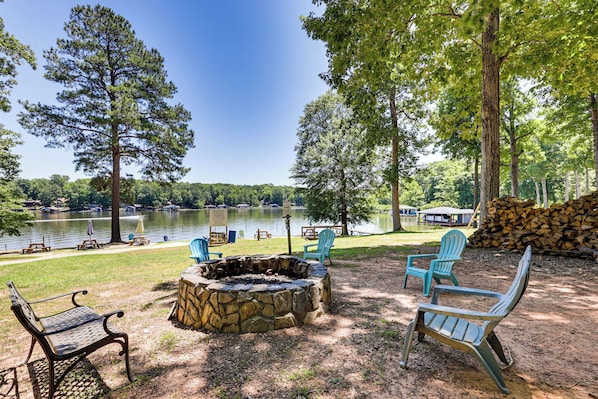 A lakeside outdoor seating area with a stone fire pit, picnic tables, and Adirondack chairs. - It's Our Pleasure! Lake Escape w/ Boat Ramp (Macon)