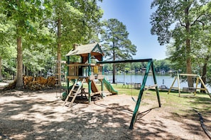 A playground with a wooden playhouse, swings, and a slide, surrounded by trees and a lake.