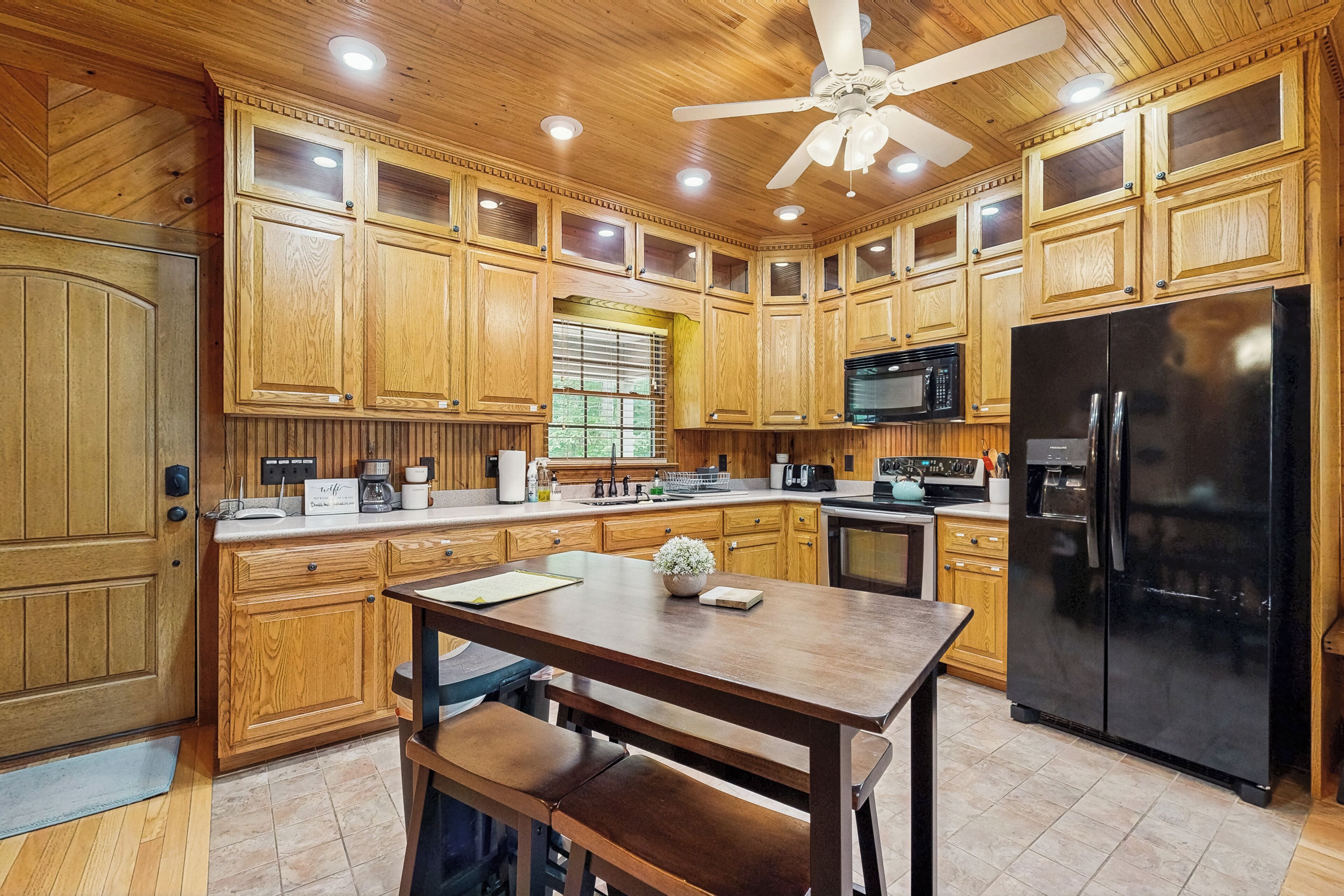 Una cocina con armarios de madera, un refrigerador negro, un microondas y un ventilador de techo.