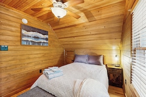 A cozy wooden cabin bedroom with a bed, a nightstand, a ceiling fan, and a painting of a snowy landscape. - Dock & Fire Pit! Lake Gaston Cabin in Macon (Macon)