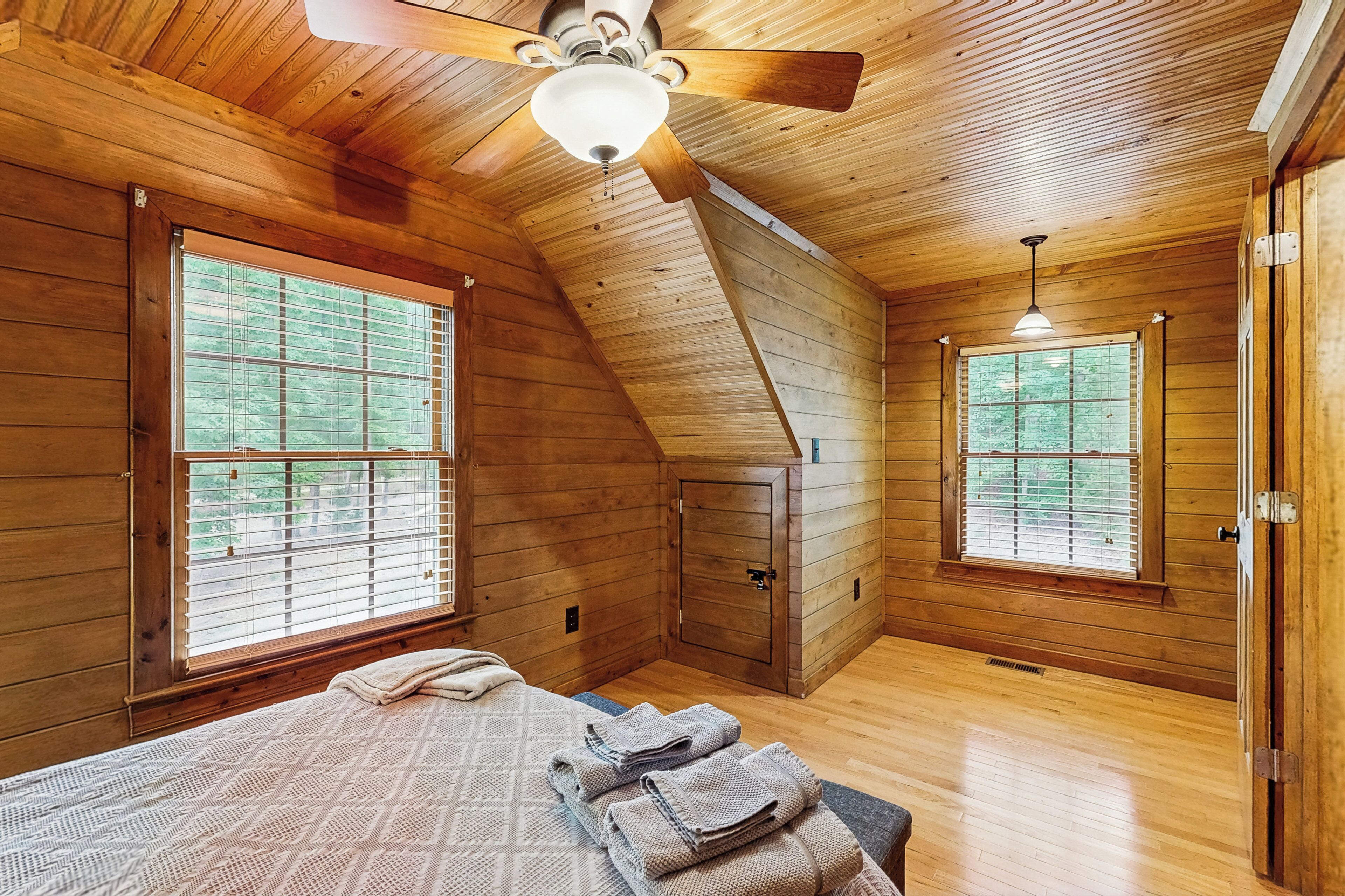A wooden cabin room with a bed, two windows, a ceiling fan, and a wooden wardrobe.