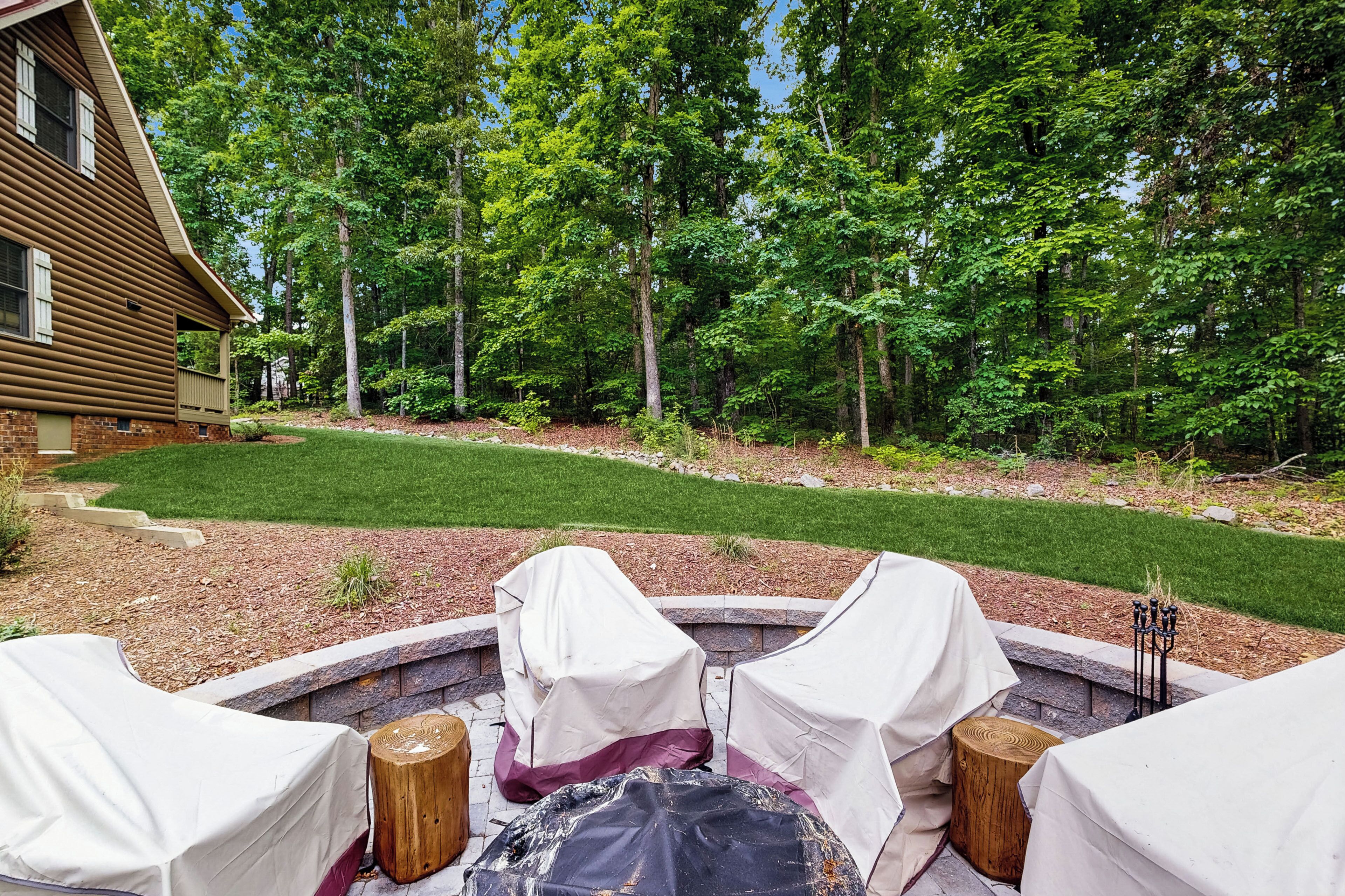 An outdoor seating area with covered chairs and a fire pit, surrounded by a well-maintained lawn and trees.