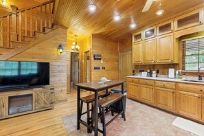 A wooden interior with a staircase, a TV, a wooden table with benches, and a kitchen area with wooden cabinets and a sink.