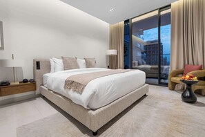 A modern bedroom with a large bed, a nightstand, a lamp, a chair with a fruit bowl, and a view of buildings through a glass door. - Arjan Studio with Pool View, Jogging Track & BBQ (Arjan)