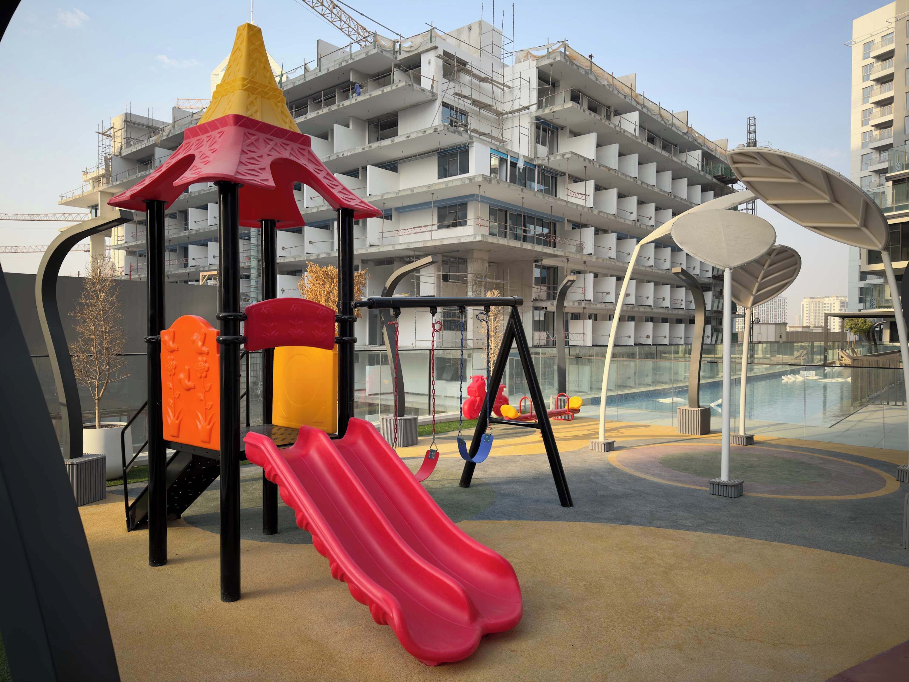 A playground with a slide and climbing structures, surrounded by a modern building under construction.