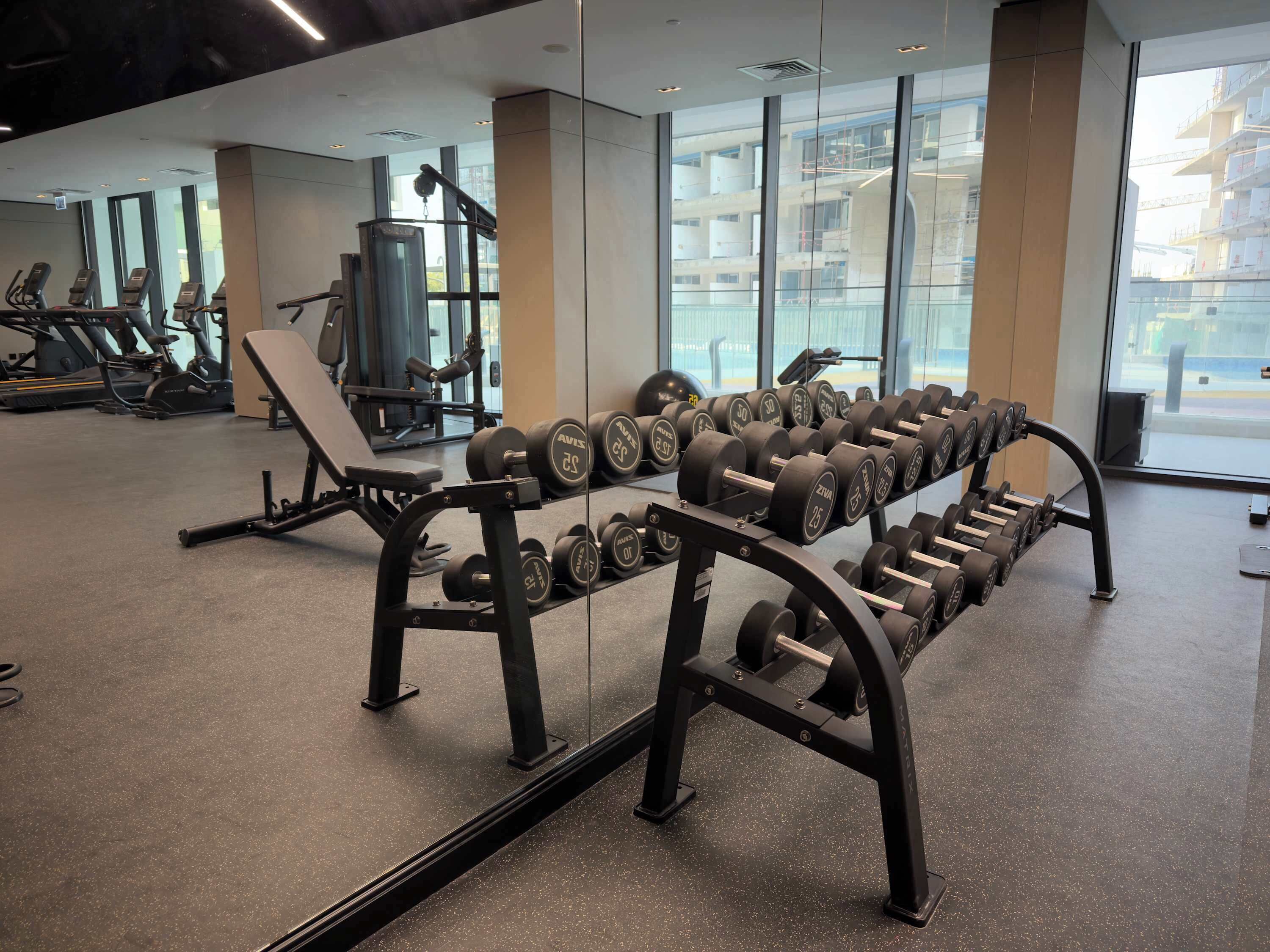 A gym with exercise equipment, including dumbbells and benches, and large windows offering a view of the outside.