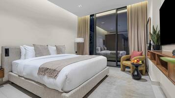A modern hotel room with a large bed, a seating area with a plant, and a view through a glass door.