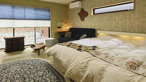Quarto com uma cama grande, uma mesinha de cabeceira, uma cadeira, uma pequena mesa e uma janela com persianas.